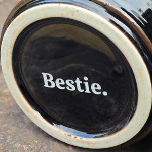 May include: A black ceramic mug featuring a white circular base rim and the word Bestie. printed in white lettering on the bottom surface. The glossy black finish covers the exterior, while the base remains unglazed with a light beige tone.