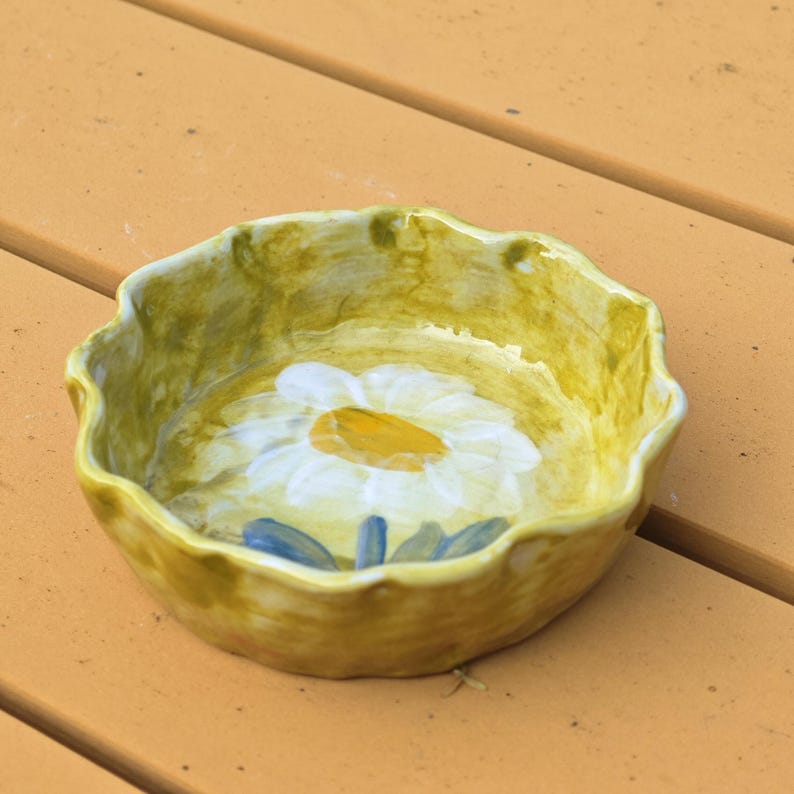 May include: A small, handmade ceramic bowl with a scalloped edge. The bowl is painted with a large white daisy with a yellow center and blue leaves against a yellow background. The bowl is approximately 6 inches in diameter.