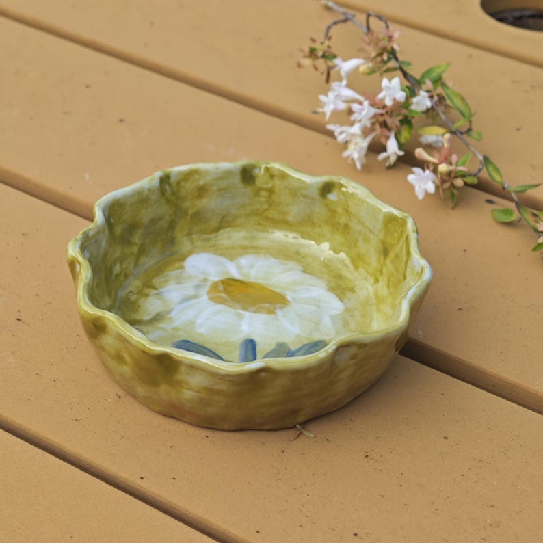 May include: A handmade ceramic bowl with a daisy design. The bowl has a scalloped edge and a yellow-green exterior. The interior features a large white daisy with a yellow center and blue accents. A decorative piece for the home.