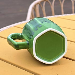 May include: A green ceramic mug with a geometric design. The mug has a hexagonal base and a matching handle. The interior is white, and the exterior has a glossy, textured finish. The mug is sitting on a yellow surface.