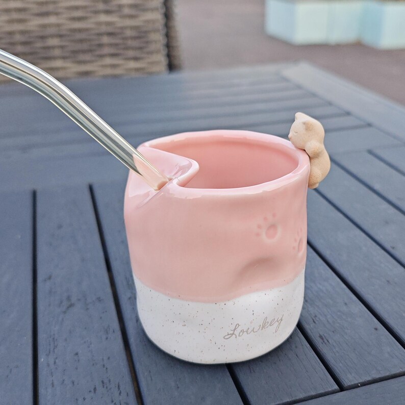 May include: A pink and white ceramic mug with a cat figurine attached to the side. The mug has paw prints and the word "Loukey" on the bottom. A silver straw is inserted into the mug.