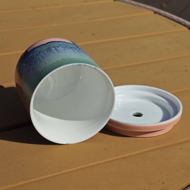 Pu&ograve; includere: Un vaso di ceramica con il suo sottovaso abbinato. Il vaso &egrave; cilindrico con un interno bianco e un esterno sfumato rosa, blu e verde. Il sottovaso &egrave; bianco con un bordo rosa e un foro di drenaggio.