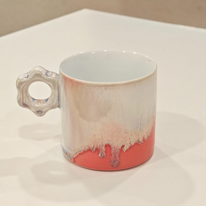 May include: A ceramic mug with a unique handle and a two-tone design. The top half is white and the bottom half is coral, with a dripped effect where the colors meet. The handle is a flower shape with a hole in the center.