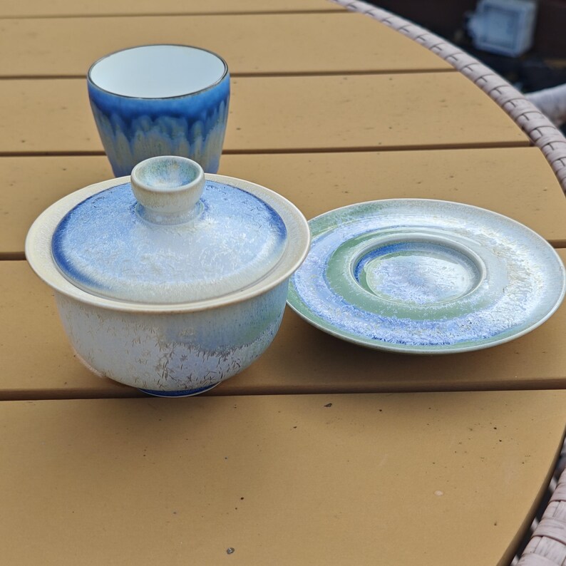 May include: A ceramic tea set with a blue and white glaze. The set includes a small lidded bowl, a cup, and a saucer. The cup and bowl have a blue interior, and the saucer has a blue and green design. The set is placed on a brown surface.