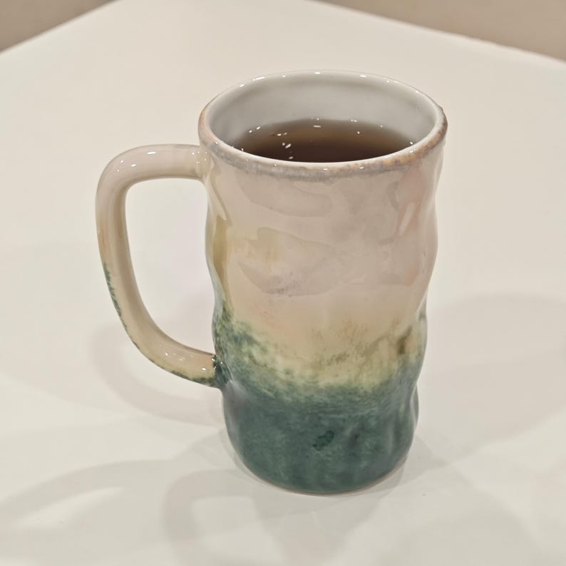 May include: A handmade ceramic mug with a unique, textured design. The mug features a gradient of colors, transitioning from a pale pink at the top to green at the base. The mug is filled with a dark liquid, likely tea or coffee.