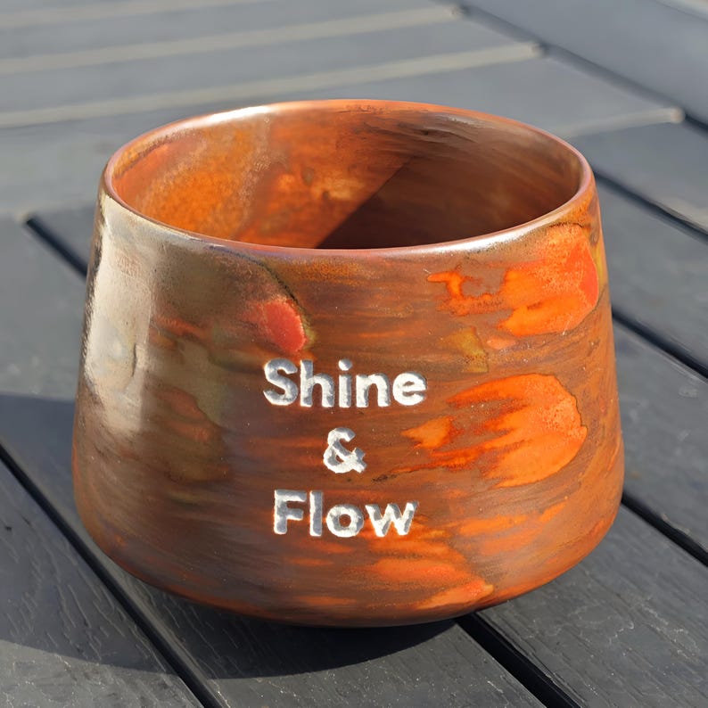 May include: A ceramic planter with a warm, earthy color palette of brown and orange. The words "Shine & Flow" are carved into the side. The planter has a wide opening and a slightly tapered shape.