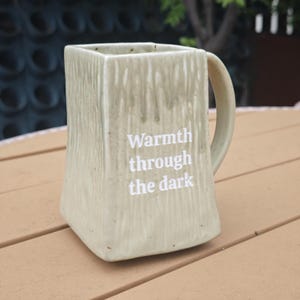 Puede incluir: Una taza de cerámica de forma cuadrada con un esmalte texturizado de color verde claro y un asa curva. La parte frontal de la taza presenta el texto impreso en blanco "Warmth through the dark" en una fuente sans-serif centrada.