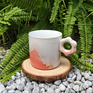 May include: A handmade ceramic mug with a unique flower-shaped handle. The mug features a white glaze with a gradient of coral and lavender hues. It sits on a wooden coaster, surrounded by small gray stones and green foliage.