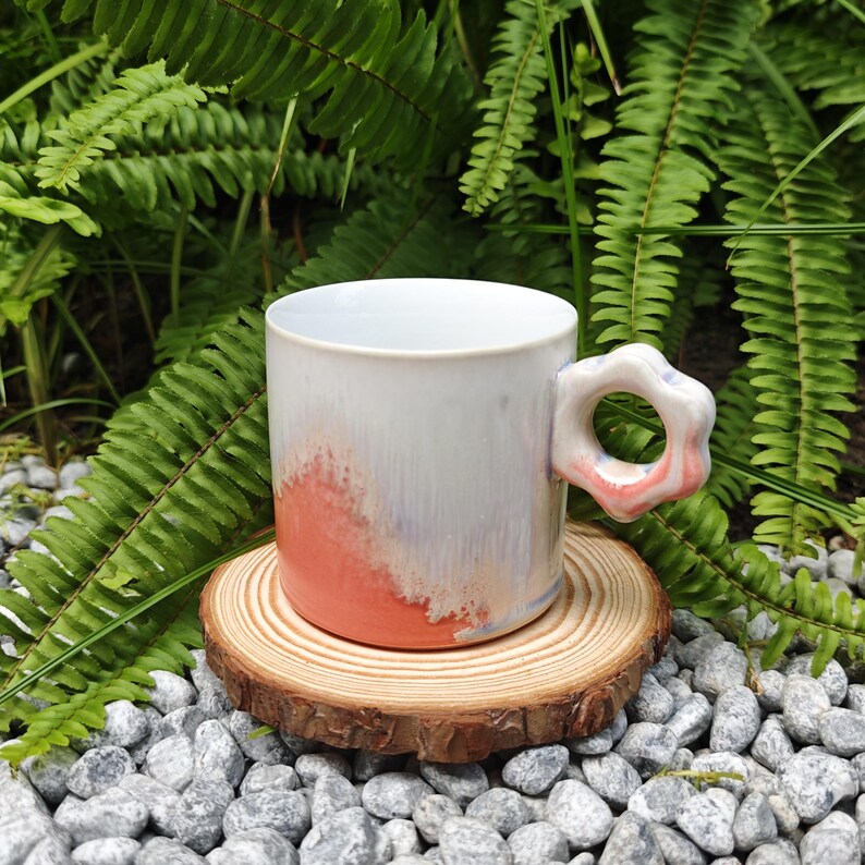 May include: A handmade ceramic mug with a unique flower-shaped handle. The mug features a white glaze with a gradient of coral and lavender hues. It sits on a wooden coaster, surrounded by small gray stones and green foliage.