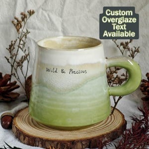 May include: A handmade ceramic mug with a green and white glaze. The mug features the text "Wild & Precious" and a green handle. The mug is on a wooden coaster, with decorative elements in the background. Custom overglaze text is available.