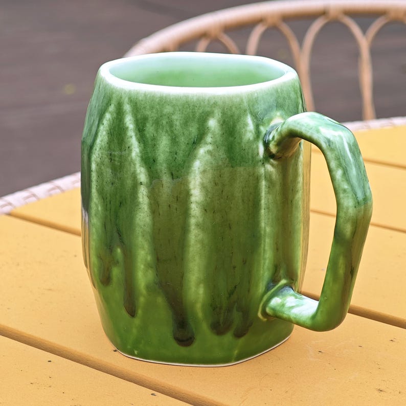 May include: A green ceramic mug with a textured exterior and a curved handle. The mug has a glossy finish with darker green streaks, suggesting a handmade or artisanal quality. The mug is sitting on a yellow surface.