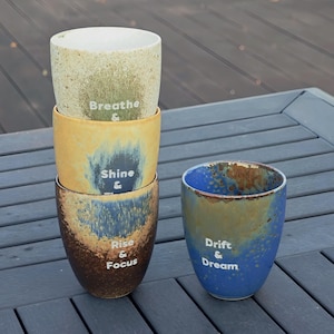 Puede incluir: Cuatro vasos de cer&aacute;mica con un acabado moteado y esmaltado en tonos beige, amarillo, azul y marr&oacute;n. Cada vaso tiene una palabra o frase inspiradora diferente en texto blanco: "Breathe", "Shine", "Rise & Focus" y "Drift & Dream".