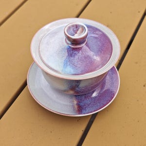 May include: A ceramic gaiwan tea set with a lid, bowl, and saucer. The set features a gradient of colors, including purple, blue, and white. The tea set is designed for brewing and serving tea. The set is sitting on a wooden surface.