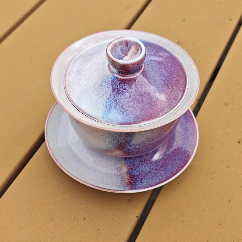 May include: A ceramic gaiwan tea set with a lid, bowl, and saucer. The set features a gradient of colors, including purple, blue, and white. The tea set is designed for brewing and serving tea. The set is sitting on a wooden surface.