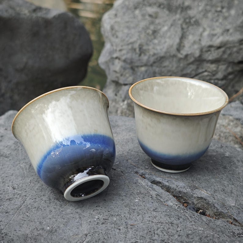 May include: Two ceramic tea cups featuring a smooth, glossy glaze that transitions from a creamy off-white at the rim to a deep, saturated blue at the base. The cups have a rounded, flared shape with a thin, light-colored rim and a dark foot ring.