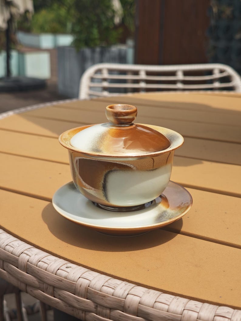 May include: A ceramic tea bowl with a matching lid and saucer, featuring a glazed finish with swirling patterns of brown, cream, and dark grey. The bowl has a wide rim and a small knob on the lid, showcasing a smooth, reflective surface throughout.