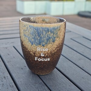 Puede incluir: Un vaso de cer&aacute;mica con un esmalte moteado marr&oacute;n, beige y azul. Las palabras "Rise & Focus" est&aacute;n impresas en blanco en la parte delantera. El vaso es cil&iacute;ndrico con una parte superior ligeramente ensanchada.