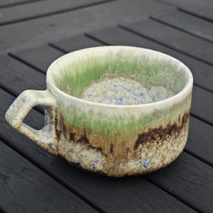 May include: A handmade ceramic mug with a unique, textured glaze. The mug features a handle and a wide, shallow bowl. The colors include shades of green, brown, and blue, creating a natural, earthy aesthetic. The mug is suitable for coffee, tea, or soup.