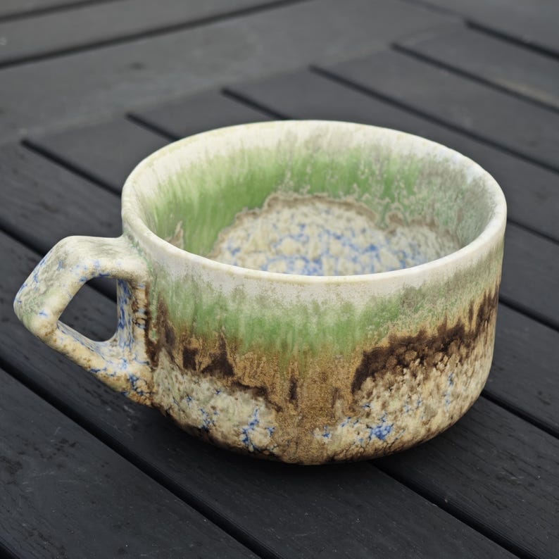May include: A handmade ceramic mug with a unique, textured glaze. The mug features a handle and a wide, shallow bowl. The colors include shades of green, brown, and blue, creating a natural, earthy aesthetic. The mug is suitable for coffee, tea, or soup.