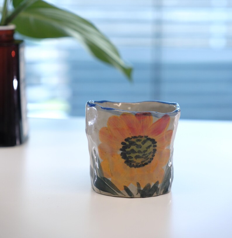 May include: A small, handmade ceramic cup with a rough, textured exterior. The cup features a vibrant, hand-painted sunflower design in shades of yellow, orange, and green. The rim is outlined with a blue band. The cup is sitting on a white surface.