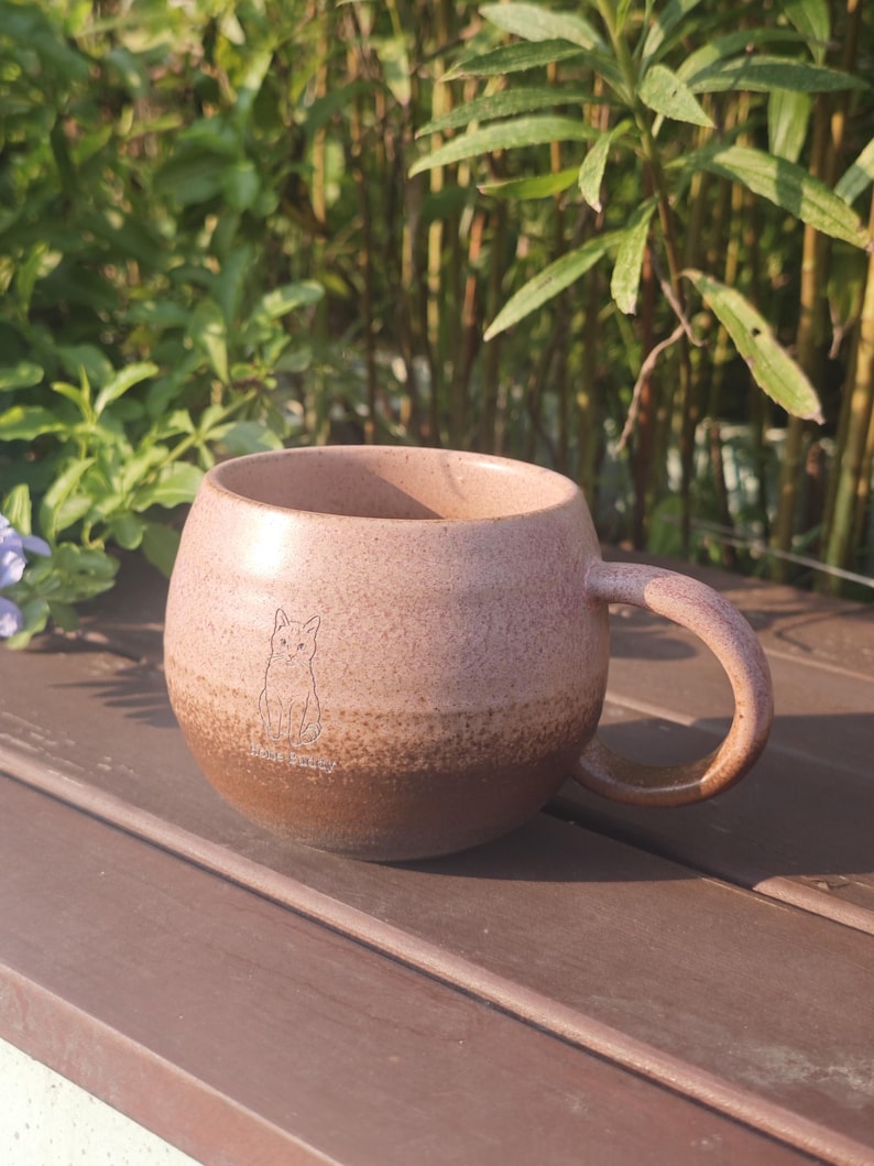 Może przedstawiać: Okrągły ceramiczny kubek z nakrapianym wykończeniem, charakteryzujący się gradientowym przejściem kolor&oacute;w od jasnego, przybrudzonego r&oacute;żu u g&oacute;ry do głębokiego brązu u podstawy. Z przodu znajduje się mały, prosty rysunek kota z tekstem poniżej.