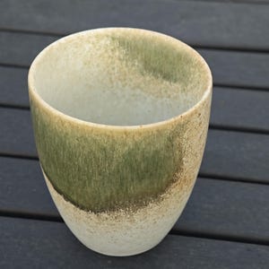 Puede incluir: Una taza de cer&aacute;mica hecha a mano con un esmalte &uacute;nico. La taza presenta una mezcla de tonos verdes, beige y blancos, creando una apariencia texturizada y org&aacute;nica. El dise&ntilde;o de la taza es simple, con una base redondeada y un borde ligeramente ensanchado.