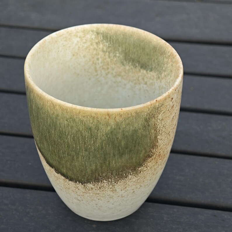 Puede incluir: Una taza de cer&aacute;mica hecha a mano con un esmalte &uacute;nico. La taza presenta una mezcla de tonos verdes, beige y blancos, creando una apariencia texturizada y org&aacute;nica. El dise&ntilde;o de la taza es simple, con una base redondeada y un borde ligeramente ensanchado.