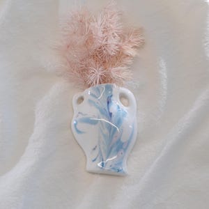 Op de afbeelding: Een kleine, witte keramische vaas met een gemarmerd blauw ontwerp en twee handvatten. De vaas bevat een bosje lichtroze gedroogde bloemen. De vaas staat op een wit, getextureerd oppervlak.