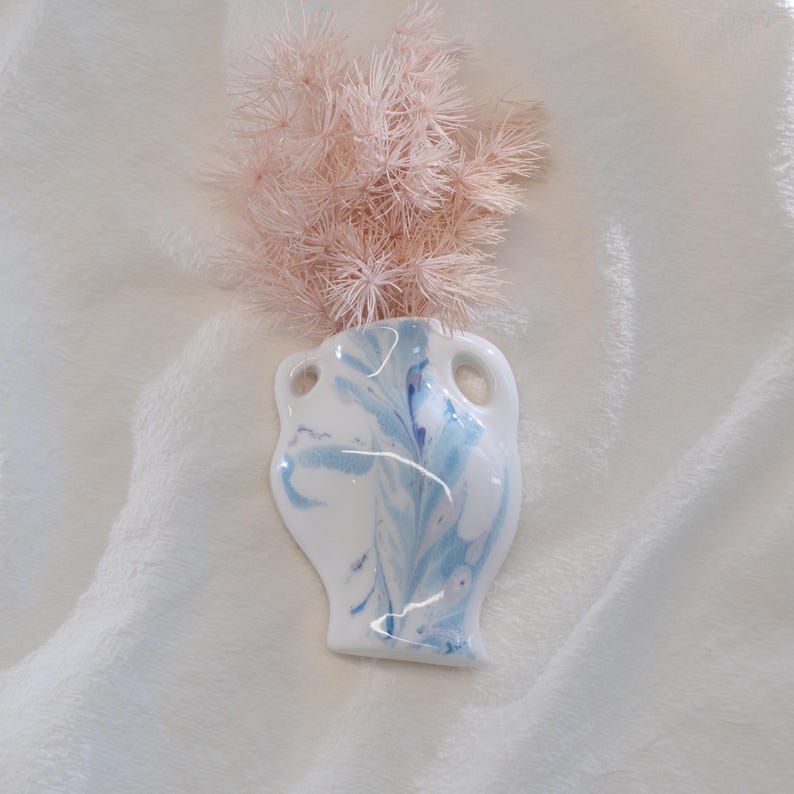 Op de afbeelding: Een kleine, witte keramische vaas met een gemarmerd blauw ontwerp en twee handvatten. De vaas bevat een bosje lichtroze gedroogde bloemen. De vaas staat op een wit, getextureerd oppervlak.