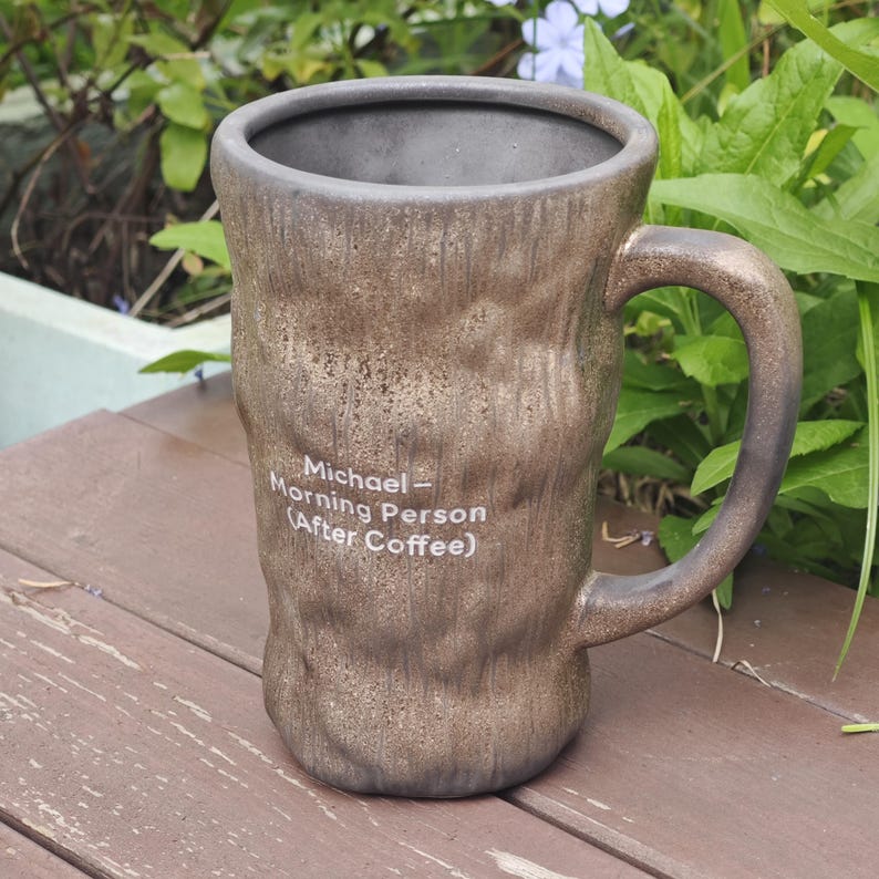 Puede incluir: Una taza alta de cer&aacute;mica marr&oacute;n con textura, acabado oscuro irregular y un asa resistente. En la parte frontal tiene impreso en blanco el texto: Michael - Morning Person (After Coffee). El interior de la taza es de color gris oscuro liso.