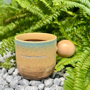 May include: A ceramic mug with a light brown base and a blue-green ombre top, filled with a dark liquid. The mug has a wooden ball handle and the words "Feeling Peachy" are printed on the side. The mug is surrounded by green ferns and gray stones.