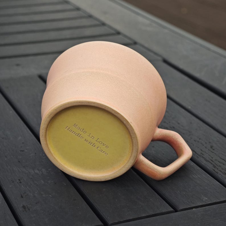 May include: A peach-colored ceramic mug with a rounded body and a curved handle. The base is yellow and features the text "Made in Love Handle with Care". The mug is resting on a dark wooden surface.