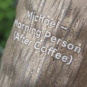 Puede incluir: Una superficie texturizada de grano de madera marr&oacute;n claro con el texto blanco grabado: Michael - Morning Person (After Coffee). La superficie tiene un aspecto natural y org&aacute;nico con patrones de grano verticales y un acabado mate poroso.