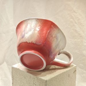 May include: A handmade ceramic mug with a wide, rounded shape. The mug features a gradient of colors, transitioning from a deep red at the base to a pearlescent white and purple at the rim. It has a curved handle and a smooth, glossy finish.