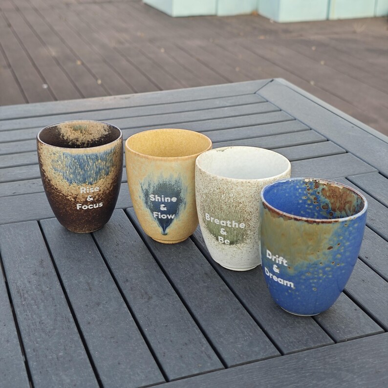Puede incluir: Cuatro tazas de cer&aacute;mica con frases inspiradoras. Las tazas tienen un acabado moteado y vidriado en tonos marr&oacute;n, azul, amarillo y blanco. Cada taza tiene una frase diferente: "Rise & Focus", "Shine & Flow", "Breathe & Be" y "Drift & Dream".