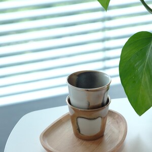 以下が含まれることがあります： 木製のトレイに重ねられた2つのセラミックカップ。カップは白で、茶色の縁と底があります。トレイは明るい茶色です。背景には緑の植物があります。