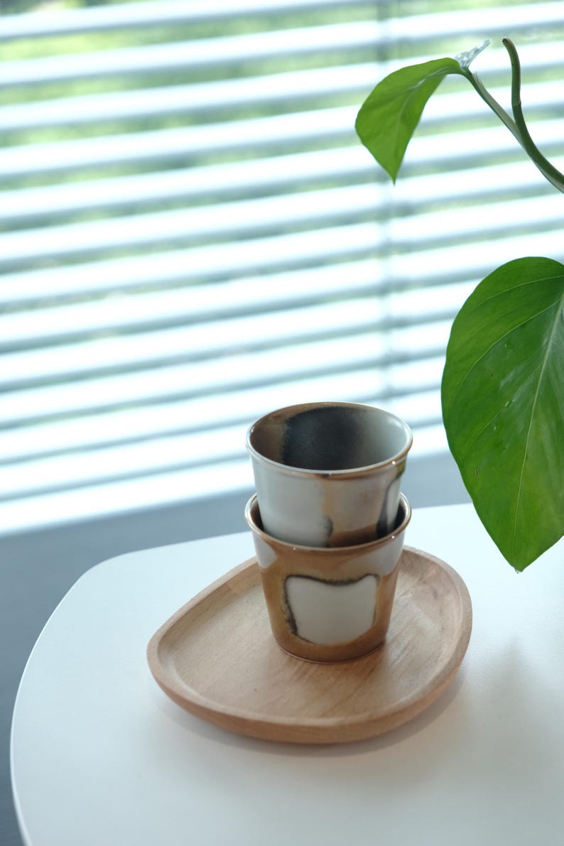 以下が含まれることがあります： 木製のトレイに重ねられた2つのセラミックカップ。カップは白で、茶色の縁と底があります。トレイは明るい茶色です。背景には緑の植物があります。