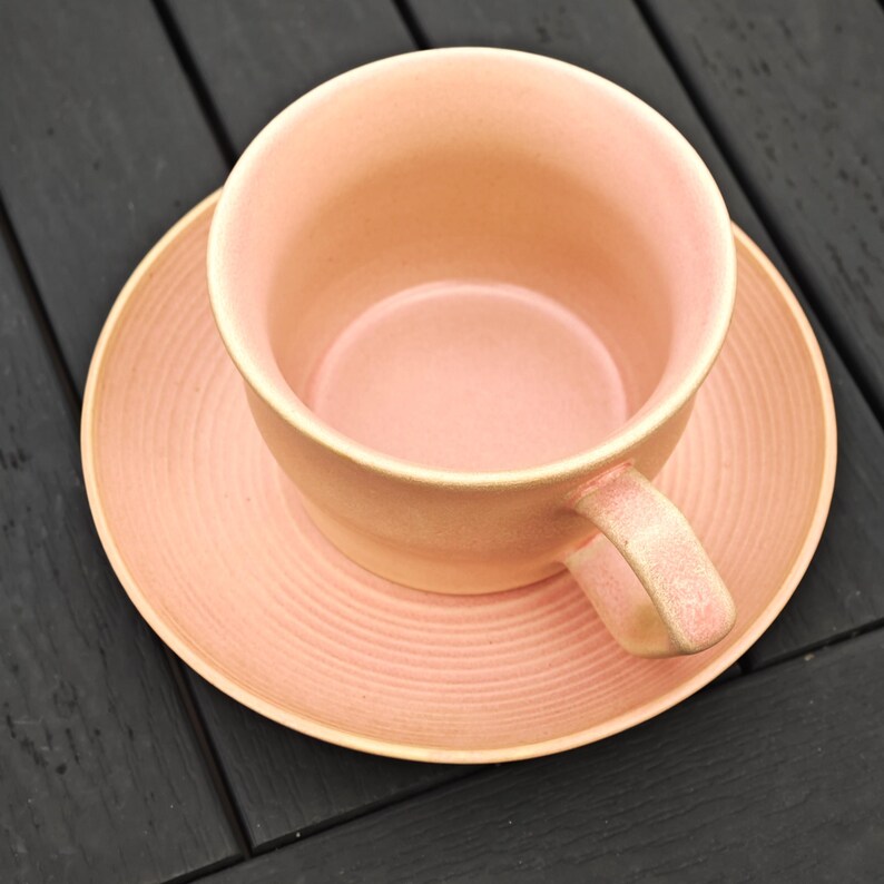 May include: A light peach-colored ceramic teacup and saucer set. The cup has a rounded shape with a handle, and the saucer has a circular design. The set is placed on a dark wooden surface.