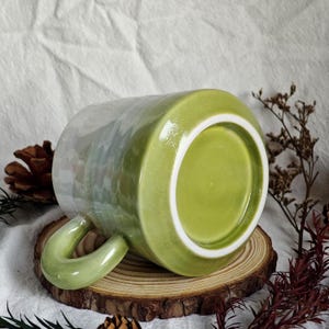 May include: A ceramic mug with a green handle and a green interior. The exterior has a pearlescent finish that transitions from green to white. The mug is resting on a wooden slice.