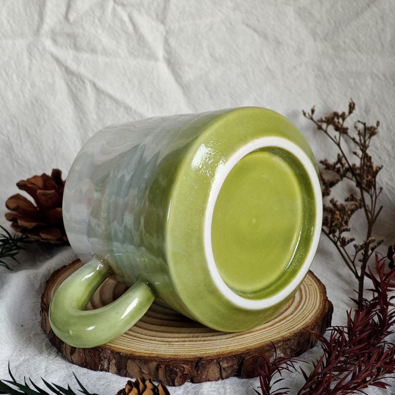 May include: A ceramic mug with a green handle and a green interior. The exterior has a pearlescent finish that transitions from green to white. The mug is resting on a wooden slice.