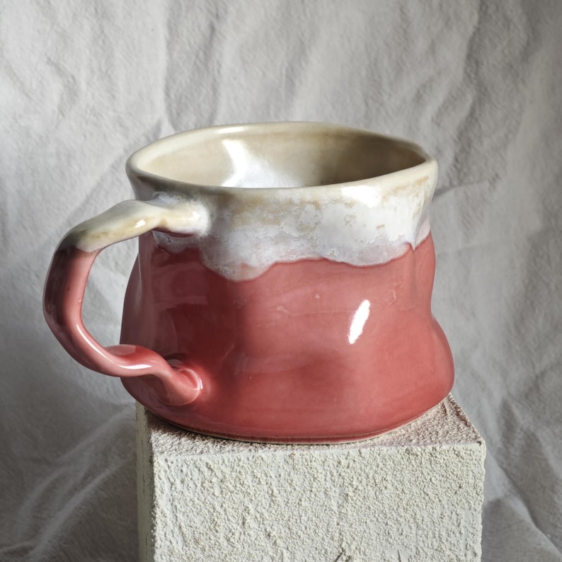 May include: A handmade ceramic mug with a pink body and a white and cream-colored rim. The mug has a curved handle and sits on a textured, square block. The mug is a unique piece of pottery.