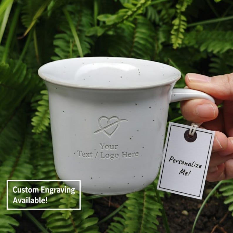 May include: A speckled white ceramic mug with a heart and arrow design, text that reads "Your Text / Logo Here," and a tag that says "Personalize Me!" The mug has a handle and is set against a green leafy background. The text "Custom Engraving Available!" is also visible.