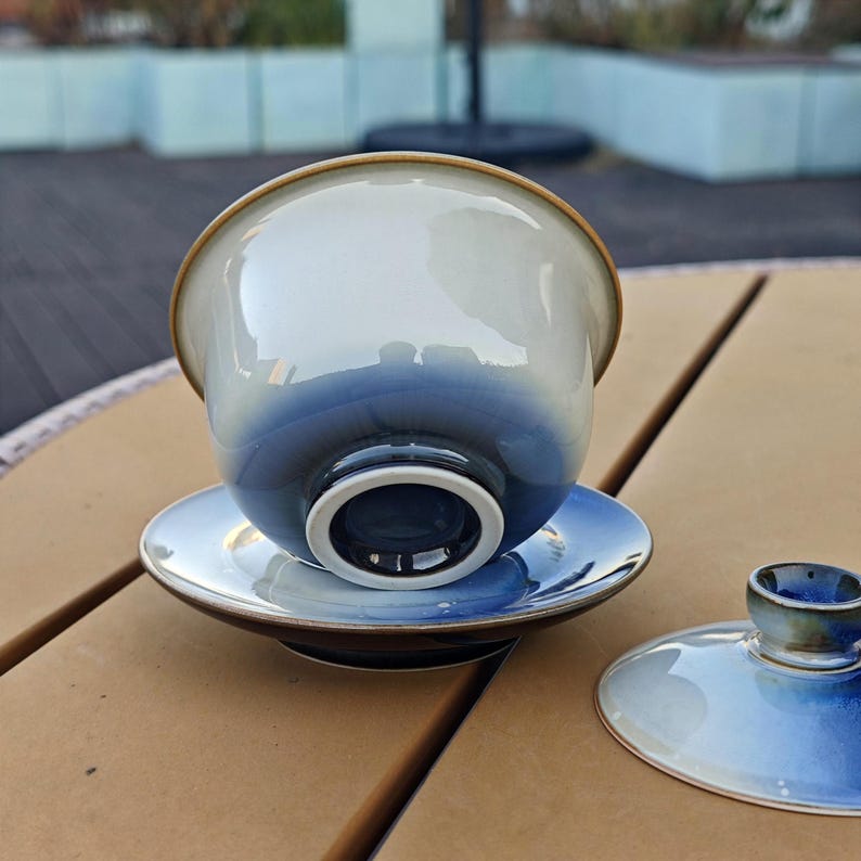 May include: A ceramic gaiwan tea set featuring a bowl, saucer, and lid with a smooth gradient finish that transitions from a deep navy blue at the base to a pale, glossy off-white at the rim, showcasing a reflective surface and a brown edge.