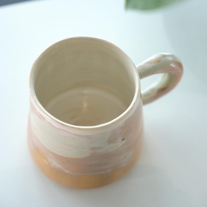 May include: A handmade ceramic mug with a rounded shape and a large handle. The mug features a color gradient, transitioning from a golden-yellow base to a pale pink and white upper section. The interior is a creamy white.
