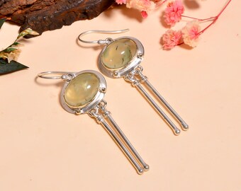 May include: A pair of silver earrings with oval, light green gemstones. Each earring features a silver frame around the gemstone, with two dangling silver bars. The earrings are displayed on a light peach surface, with pink flowers and a piece of wood in the background.