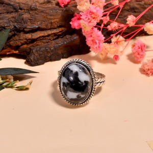 Può includere: Un anello d'argento con una pietra ovale di diaspro zebrato. La pietra presenta un motivo bianco e nero, incastonata in una lunetta a forma di corda ritorta. La fascia dell'anello ha un design strutturato. Fiori rosa e legno sullo sfondo.