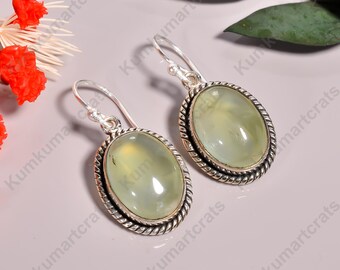 May include: A pair of oval-shaped earrings with a light green gemstone center and a silver-colored, twisted rope-like border. The earrings have a hook closure and are displayed on a light purple surface.