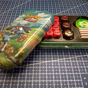 Peut inclure: Une boîte de jeu de cartes Pokémon verte et turquoise avec un motif Riolu. La boîte ouverte révèle des dés rouges, des dés noirs, des jetons verts et des compteurs blancs. La boîte est posée sur une surface quadrillée bleue.