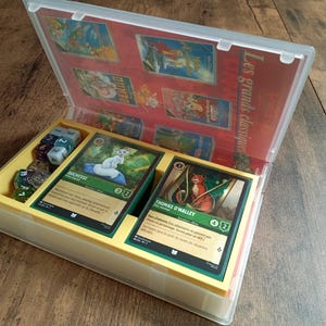 Può includere: Una custodia di plastica gialla e trasparente contenente un gioco di carte. La custodia è aperta, rivelando carte con illustrazioni e testo, dadi e uno sfondo rosso con immagini di carte aggiuntive. Il testo "Les grands classiques" è visibile.