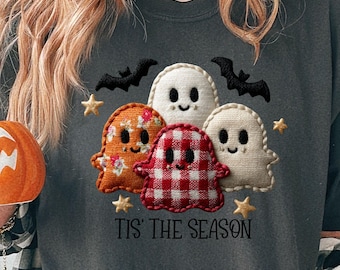 Może przedstawiać: Ciemnoszara koszulka z haftowanymi wzorami duchów w pomarańczowe kwiaty, czerwoną kratę i białe. Koszulka ma napis "Tis' the Season" i jest połączona z kubkiem w kształcie dyni. Odzież o tematyce Halloween.